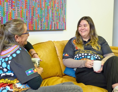 Two people sitting on a couch with mugs talking and smiling