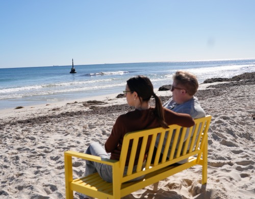 Two people sitting on a Yellow Bench facing the ocean