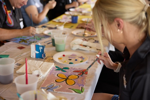 A woman painting a flower on a canvas siiting at a table with others doing artwork too.