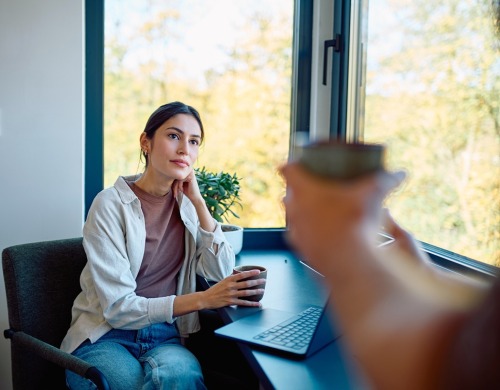 Woman having coffee, listening thoughtfully during a conversation, sitting at a window desk with a laptop