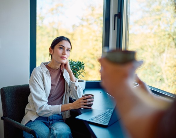 Woman having coffee, listening thoughtfully during a conversation, sitting at a window desk with a laptop